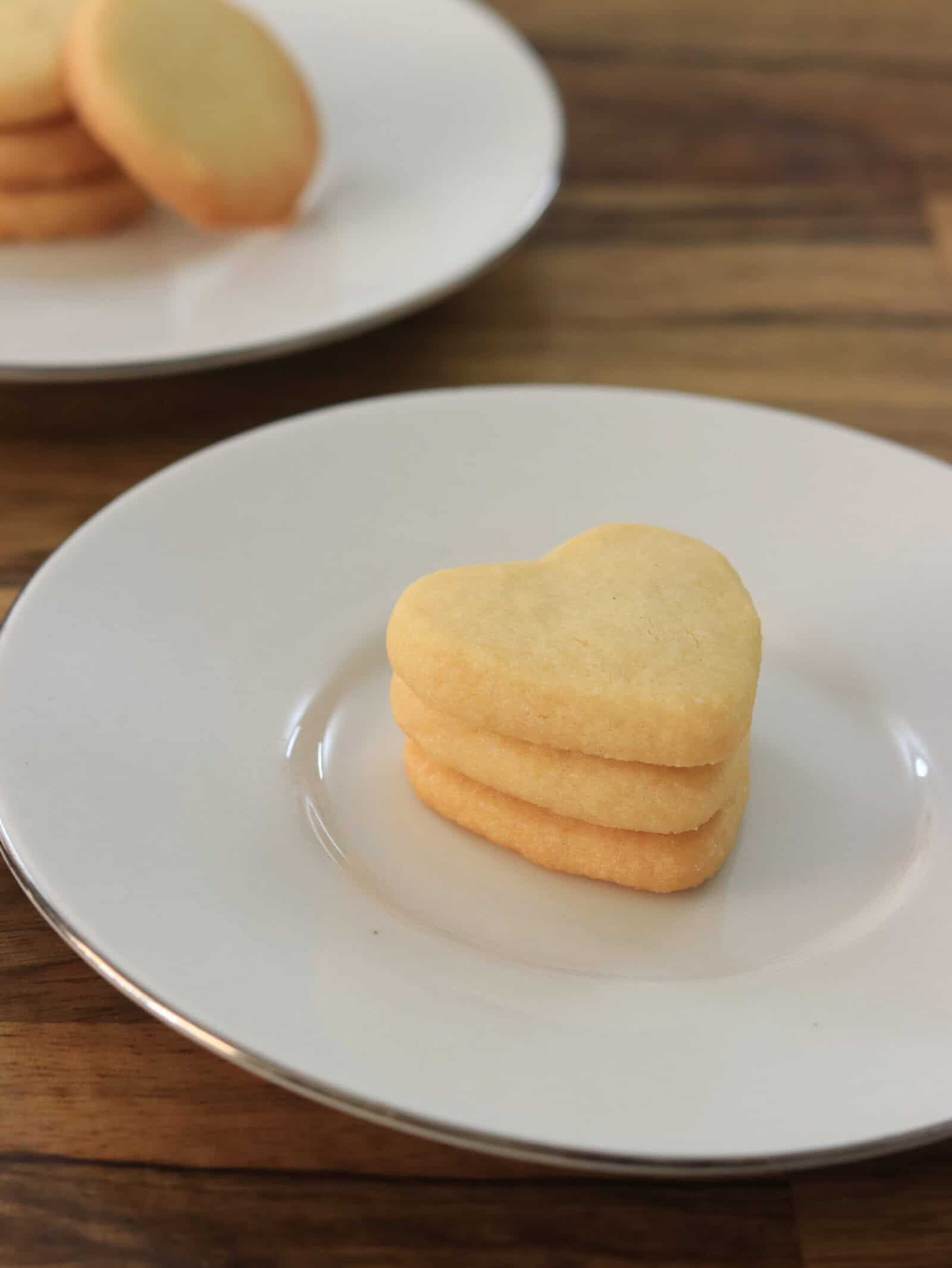 Three heart-shaped butter cookies stacked on a white plate, with more cookies on another plate in the background, all set on a wooden surface.