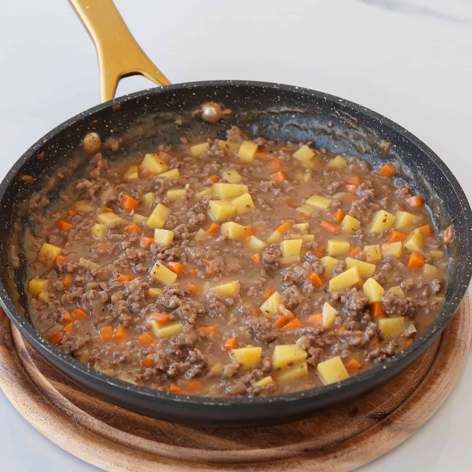 A skillet filled with a mixture of cooked ground beef, diced potatoes, and small pieces of carrots in a savory sauce, placed on a round wooden trivet.