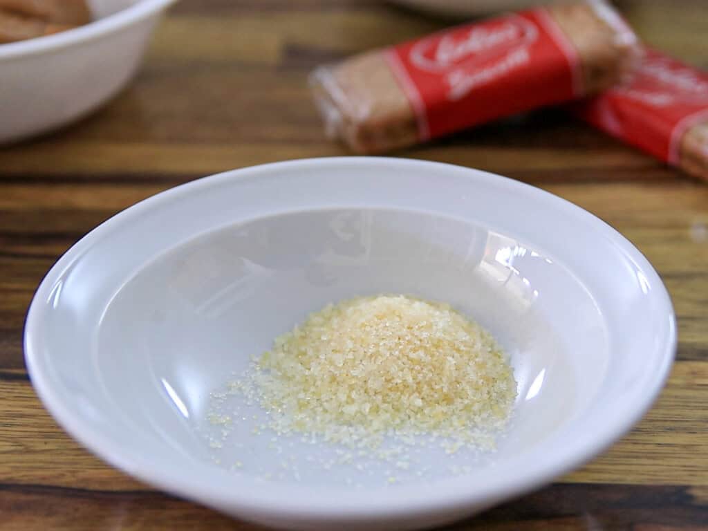 A small pile of coarse brown sugar sits in a white bowl on a wooden table, with wrapped biscuits and another bowl blurred in the background.