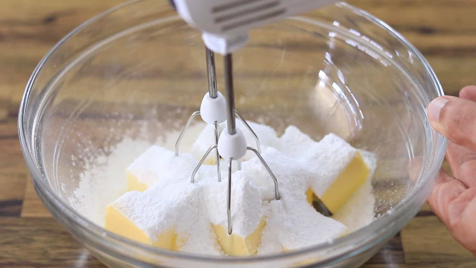A hand mixer with beaters is positioned in a glass bowl containing cubed butter and powdered sugar, ready to be mixed. The bowl is set on a wooden surface.