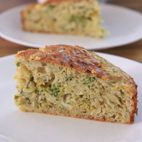 A close-up of a slice of savory cabbage cake on a white plate, with another slice in the background. The bread appears moist with visible zucchini pieces and a golden-brown crust.