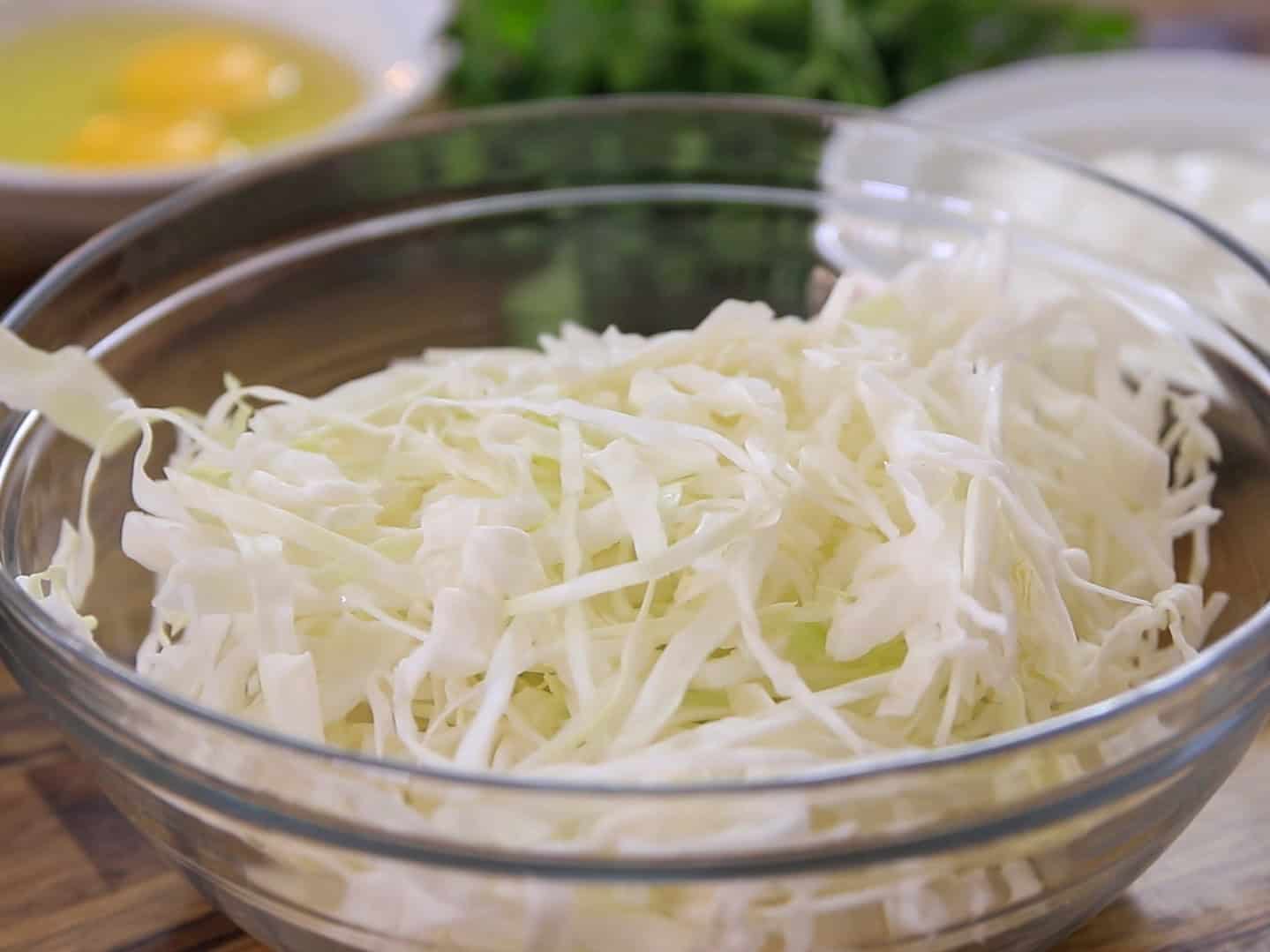A glass bowl filled with shredded cabbage sits on a wooden surface, with eggs, leafy greens, and another bowl blurred in the background.