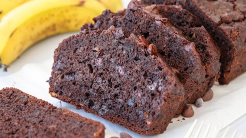 Sliced chocolate banana bread is displayed on a white surface, with chocolate chips visible in the moist, dark bread. Whole bananas are in the background.