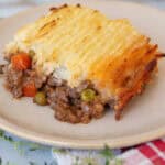 A serving of shepherd's pie on a beige plate, showing a layer of mashed potatoes on top of ground meat, peas, and carrots. The pie has a golden, crispy potato topping and is placed on a red checkered cloth.
