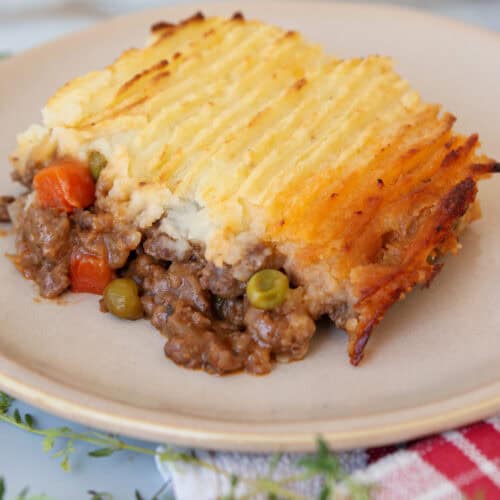 A serving of shepherd's pie on a beige plate, showing a layer of mashed potatoes on top of ground meat, peas, and carrots. The pie has a golden, crispy potato topping and is placed on a red checkered cloth.