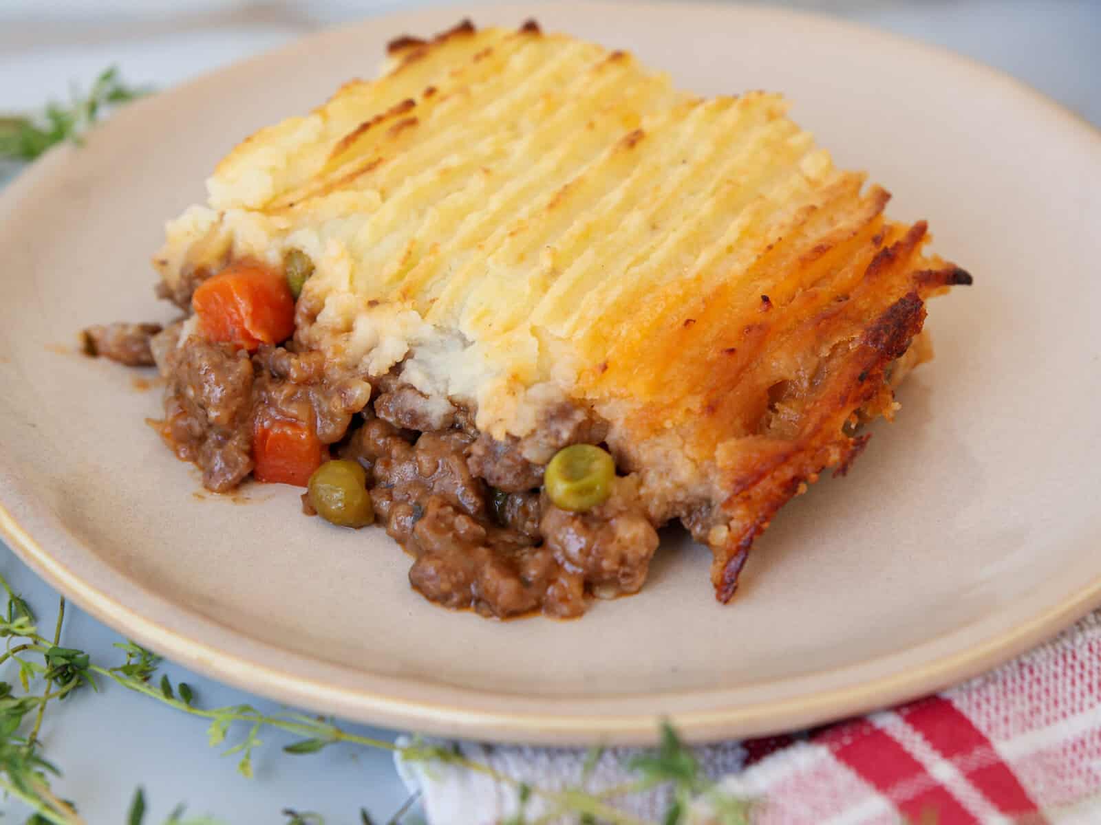 A serving of cottage pie on a beige plate, showing a layer of mashed potatoes on top of ground meat, peas, and carrots. The pie has a golden, crispy potato topping and is placed on a red checkered cloth.