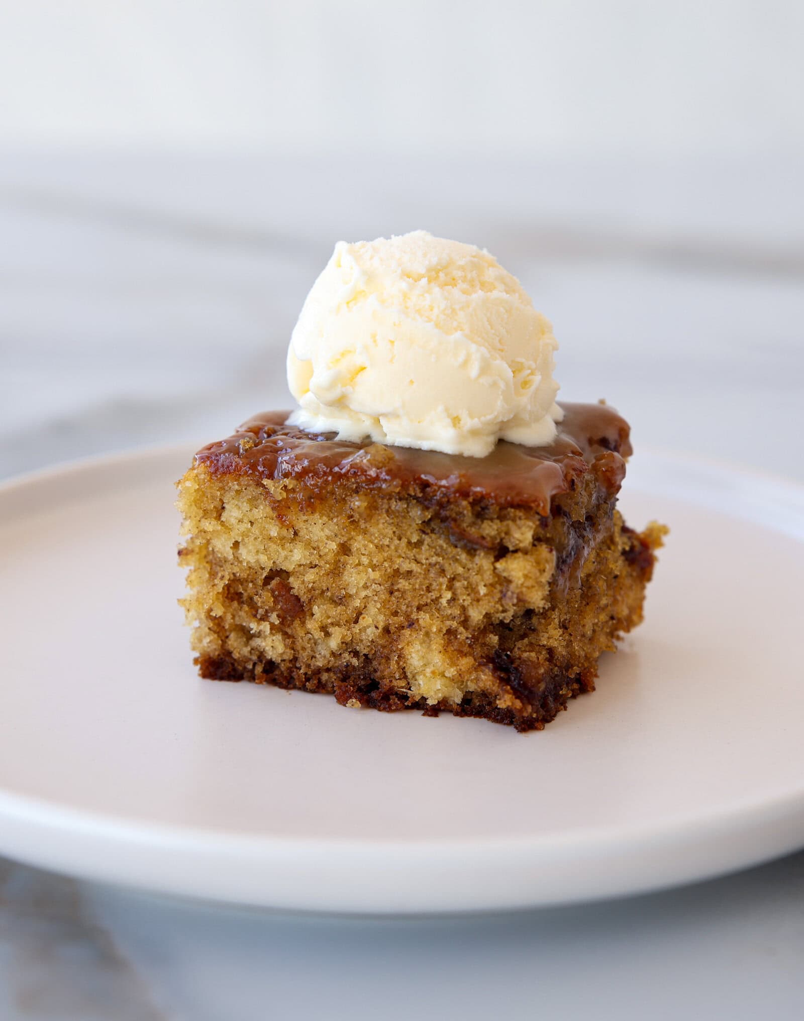 A slice of moist sticky toffee pudding topped with a scoop of vanilla ice cream on a white plate, set against a light, blurred background.
