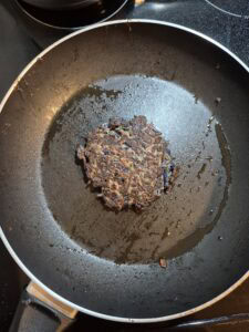 A single dark, crispy vegetable fritter is cooking in the center of a nonstick frying pan on a stovetop, with some oil visible around it.