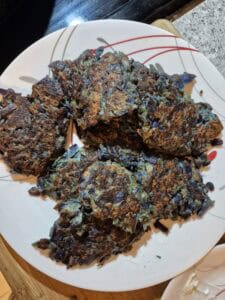 Five crispy vegetable fritters with a dark brown, slightly charred exterior rest on a white plate with red and gray stripes. The fritters feature visible shreds of purple cabbage and green herbs.