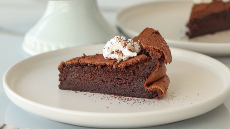 A slice of rich chocolate cake topped with whipped cream and chocolate shavings, served on a white plate.