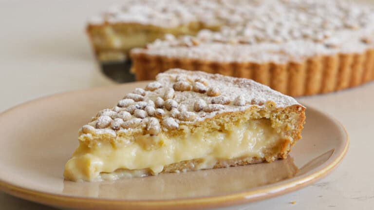 A slice of creamy custard tart topped with powdered sugar sits on a tan plate in front of the rest of the tart, which is also dusted with powdered sugar.