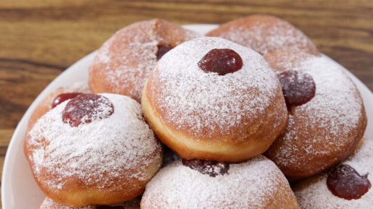 A plate of jelly-filled doughnuts dusted with powdered sugar, each topped with a dollop of red jelly, on a wooden surface.