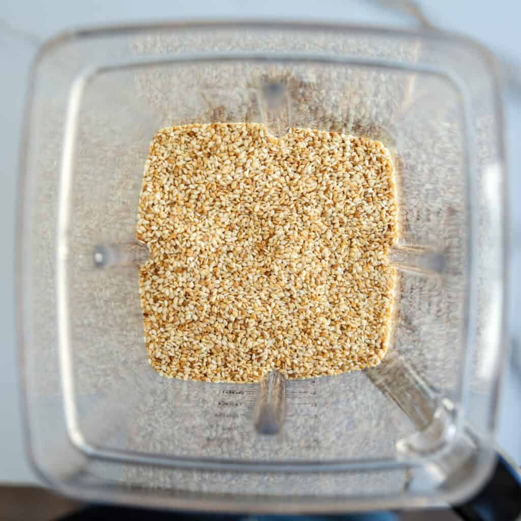 Top view of a blender containing a generous amount of raw sesame seeds, ready to be blended, with the clear plastic sides and blending blades of the container visible.