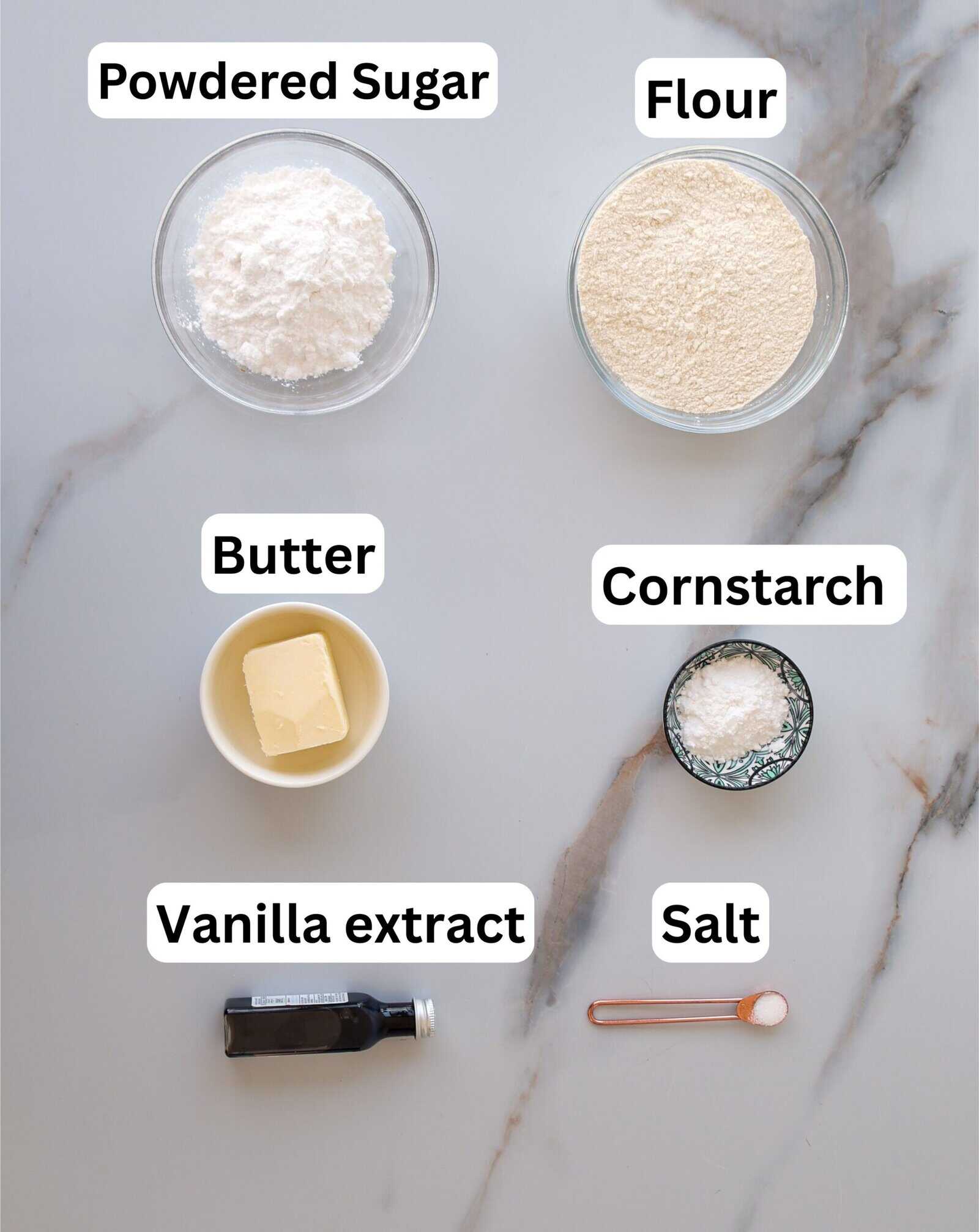Six baking ingredients on a marble surface: powdered sugar, flour, butter, cornstarch, vanilla extract in a bottle, and salt measured in a small spoon, each labeled with text.