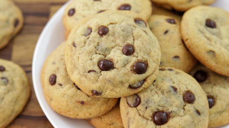 A close-up of several cchewy hocolate chip cookies stacked on a white plate, with more cookies visible in the background on a wooden surface.