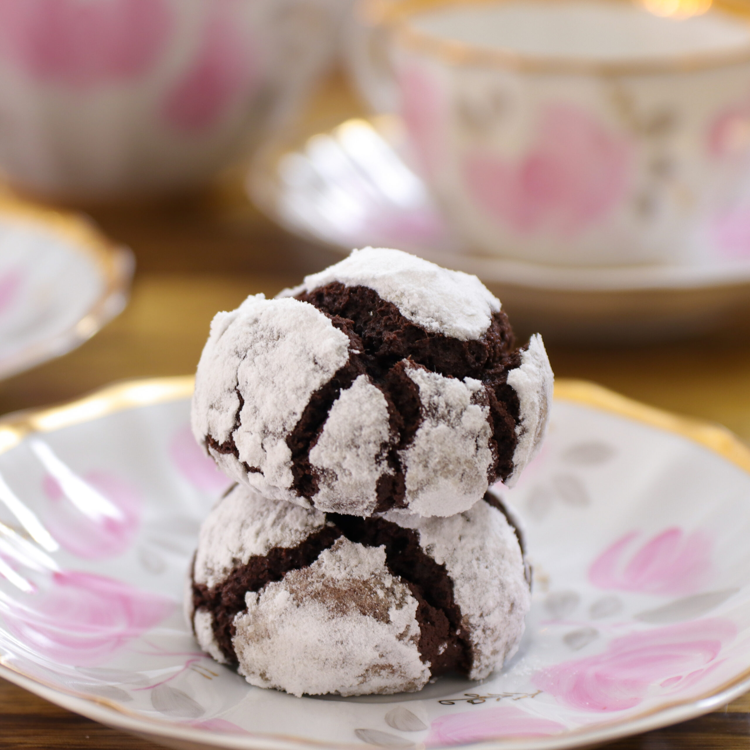 Two chocolate crinkle cookies dusted with powdered sugar are stacked on a decorative plate with pink floral patterns; teacups and matching plates are visible in the blurred background.