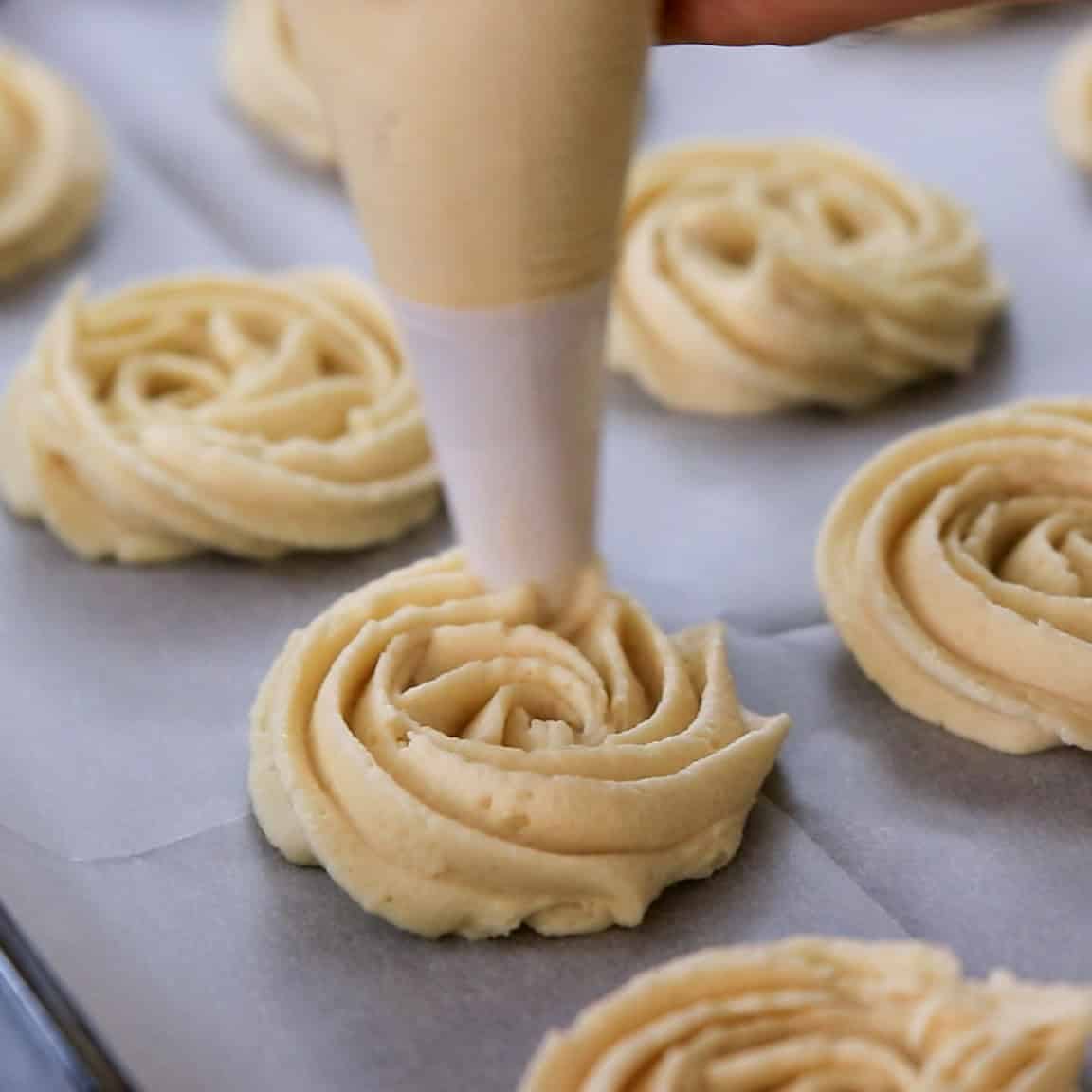 A hand uses a piping bag to pipe swirls of cookie dough onto a parchment-lined baking sheet, with several unbaked cookies visible nearby.