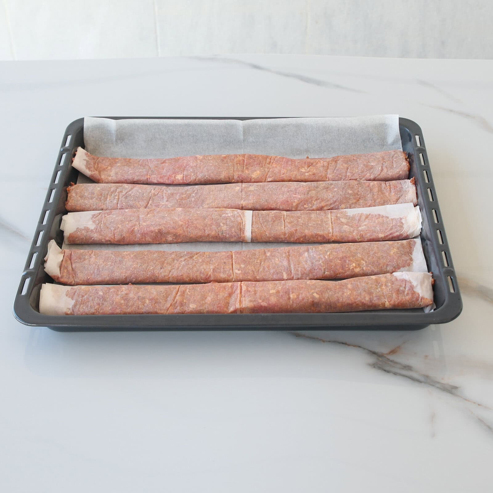 A baking tray lined with parchment paper holds six long, uncooked meat kebabs evenly spaced in a row, ready for cooking, on a white marble countertop.