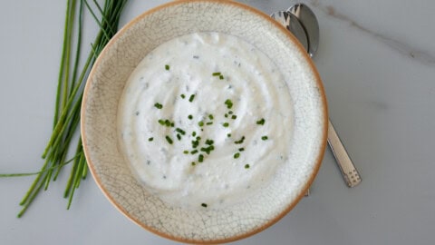 A bowl of creamy white dip garnished with chopped chives sits on a white surface, with fresh chives and two spoons beside it.