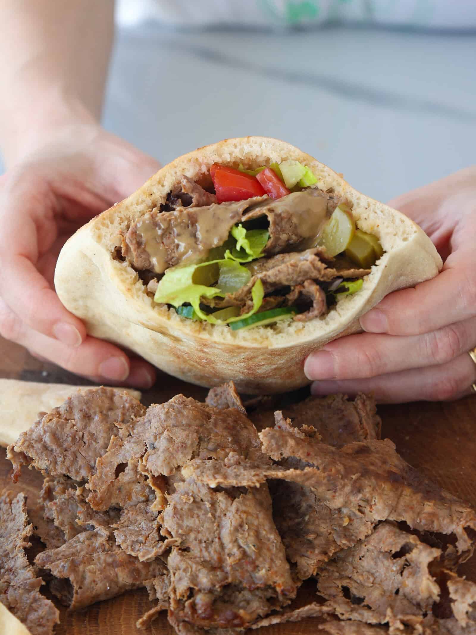 A person holds a pita filled with sliced doner beef, lettuce, pickles, and tomatoes. More slices of beef are scattered on a wooden surface below the sandwich.