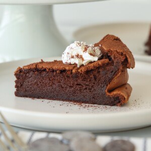 A slice of rich, flourless chocolate cake topped with a dollop of whipped cream and cocoa powder sits on a white plate. The cake has a dense, fudgy texture with slightly cracked edges.