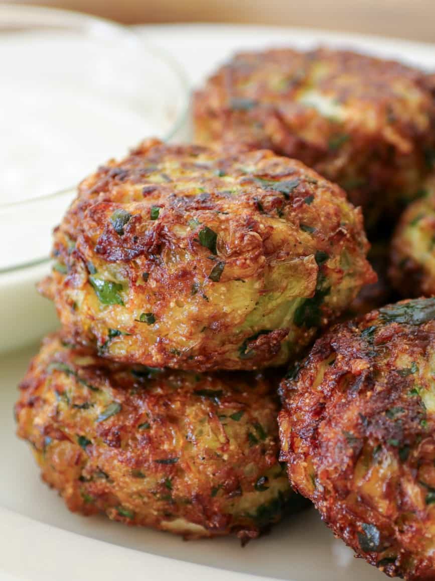 Three golden-brown, crispy cabbage-vegetable fritters stacked on a white plate, garnished with chopped herbs, with a bowl of white dipping sauce in the background.