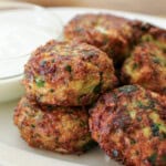 A plate of golden-brown cabbage fritters with chopped herbs, served beside a small glass bowl of white dipping sauce.