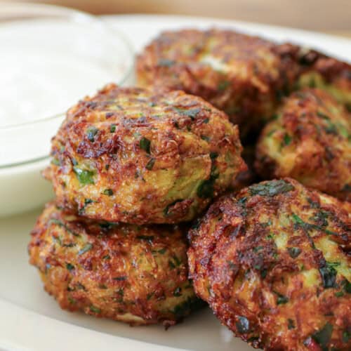 A plate of golden-brown cabbage fritters with chopped herbs, served beside a small glass bowl of white dipping sauce.