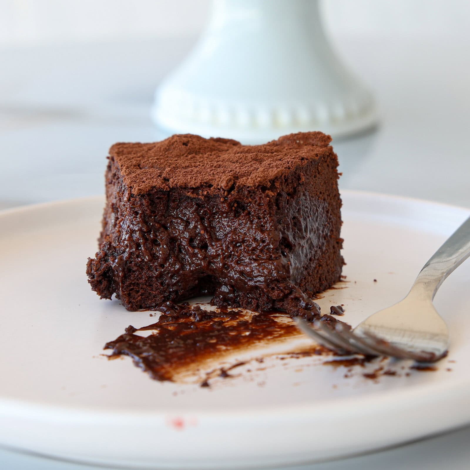 A slice of rich, moist chocolate cake with a fudgy texture sits on a white plate. A fork rests beside it, and crumbs and melted chocolate are smeared on the plate.