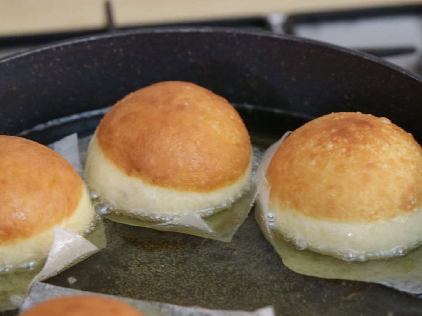 Three round dough balls are frying in hot oil in a pan, each sitting on a small piece of parchment paper. The tops are golden brown while the bottom edges are still light in color.