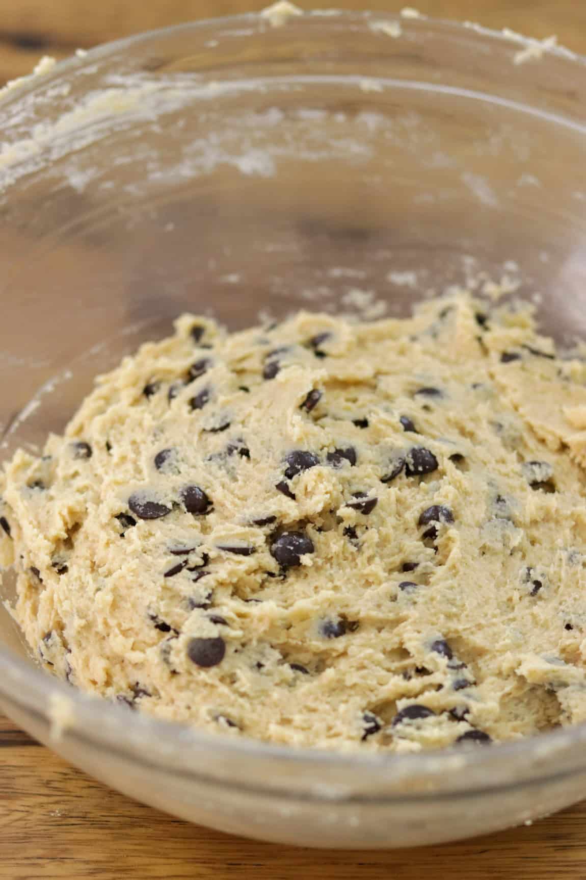 A clear glass bowl filled with chocolate chip cookie dough, mixed and ready for baking, sits on a wooden surface. The dough is creamy with visible chocolate chips throughout.