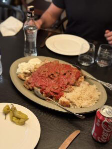 A large platter of rice, grilled meat topped with tomato sauce, pita bread, and yogurt is placed on a table set with plates, glasses, a bottled drink, and a can of Coca-Cola. A few peppers are on a side plate.