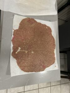 A large, thin sheet of raw ground meat mixture is spread out on a piece of parchment paper atop a gray countertop, with tiled flooring visible below.