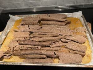 Slices of cooked, seasoned meat are spread out on a baking sheet lined with parchment paper, with some juices pooling around the edges. The background shows a tiled backsplash and part of a stovetop.