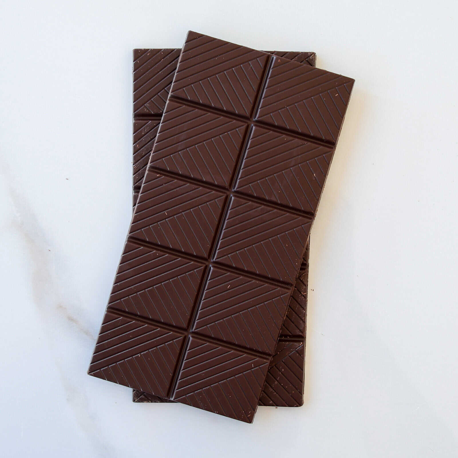 Two dark chocolate bars with geometric patterns are stacked on a white surface, with one bar slightly overlapping the other.