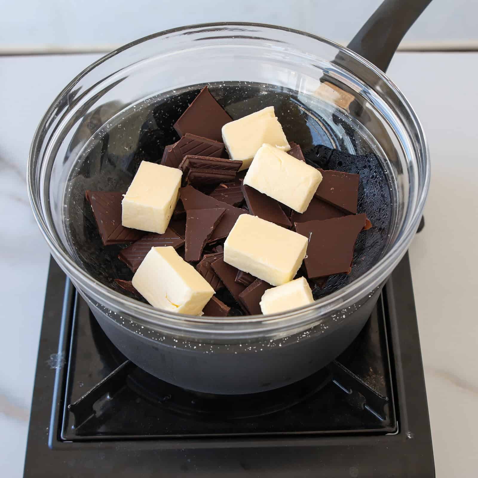 A glass bowl with chunks of dark chocolate and butter sits on top of a saucepan over a stove, showing a double boiler setup for melting the ingredients.