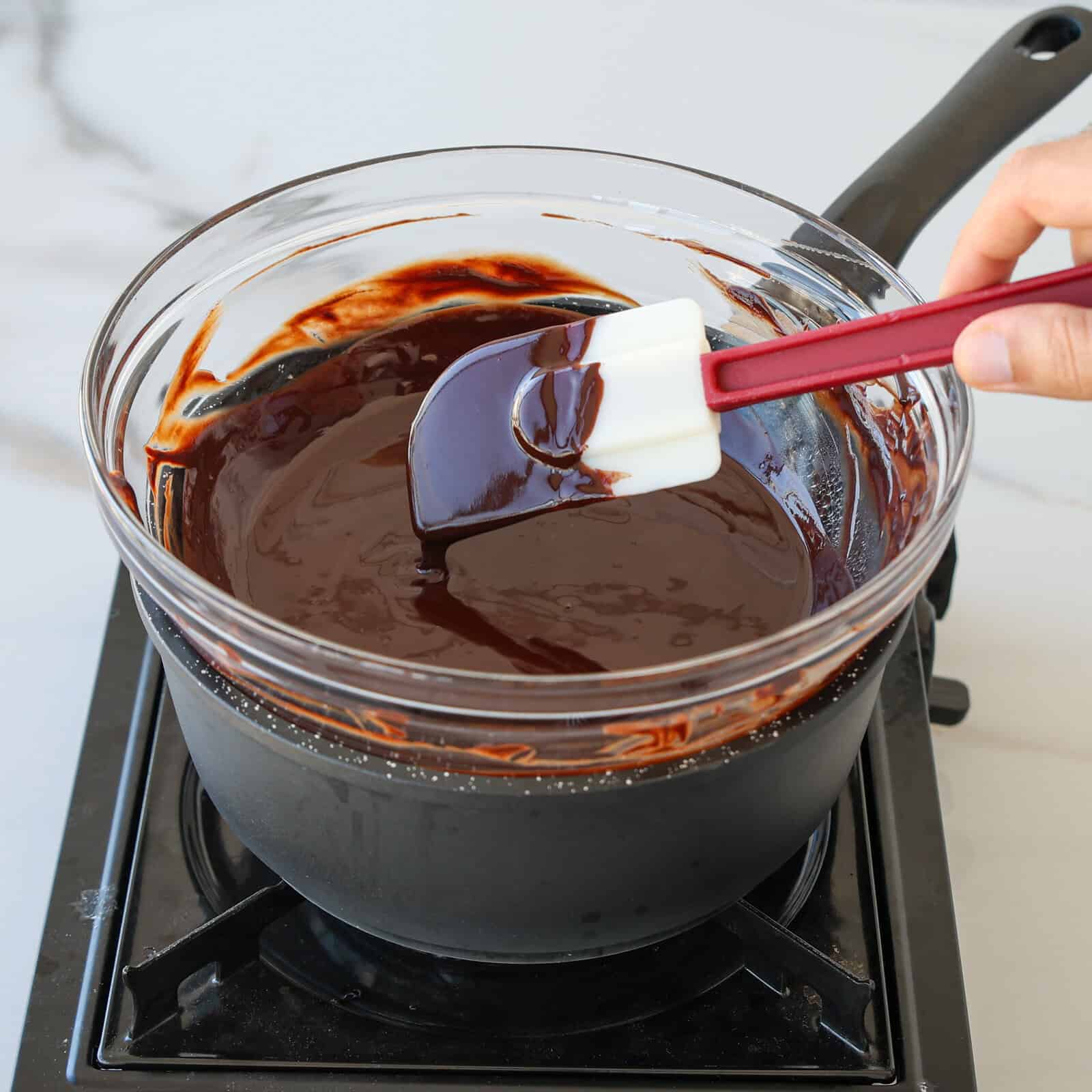 A hand uses a spatula to stir melted chocolate in a glass bowl set over a pot of simmering water on a stovetop, creating a double boiler.