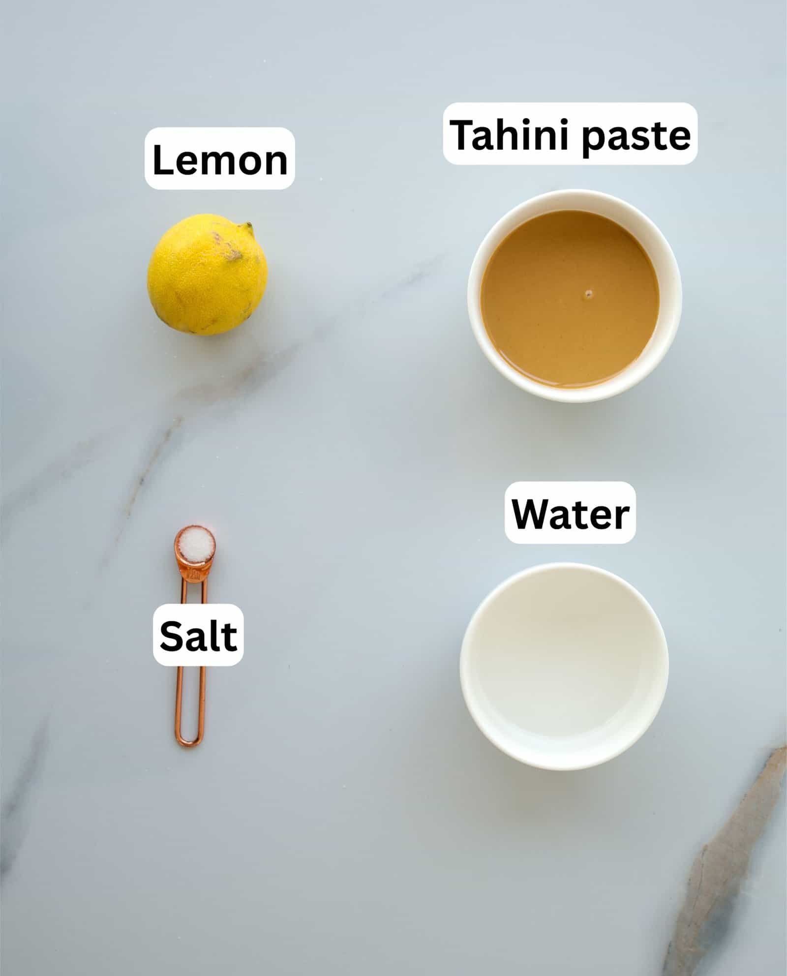 A lemon, a bowl of tahini paste, a bowl of water, and a measuring spoon of salt are arranged on a light-colored surface, each labeled with its name.