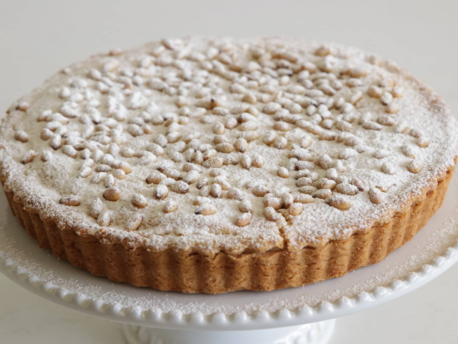 A round Italian tart (torta della nonna) with a golden crust, dusted with powdered sugar and topped with pine nuts, displayed on a white decorative cake stand.