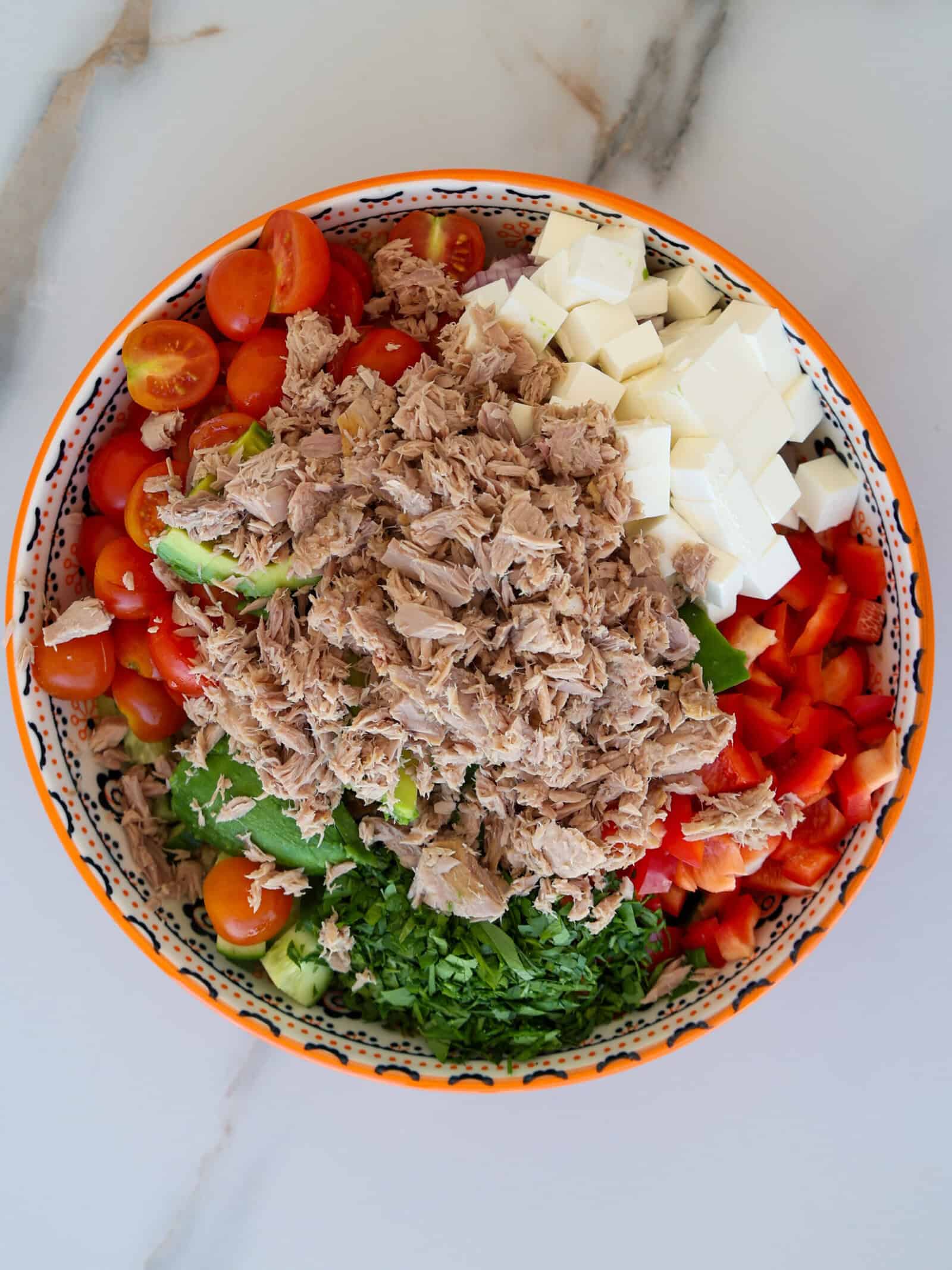 A colorful salad bowl filled with chunks of tuna, halved cherry tomatoes, diced white cheese, chopped red bell pepper, green leafy vegetables, and fresh chopped herbs, arranged on a marble surface.