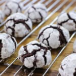 Chocolate crinkle cookies covered in powdered sugar are cooling on a metal rack over a wooden surface. The cookies have a cracked appearance with dark chocolate showing through the white sugar.