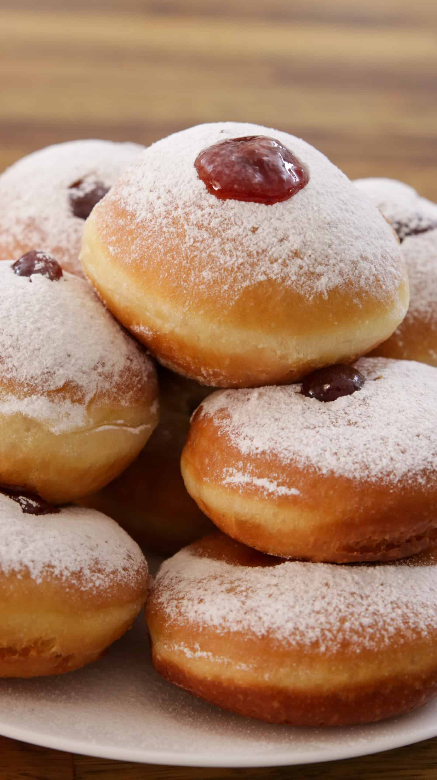 A plate stacked with powdered sugar-covered jelly donuts, each filled with red jam and topped with a dusting of sugar, on a wooden surface.