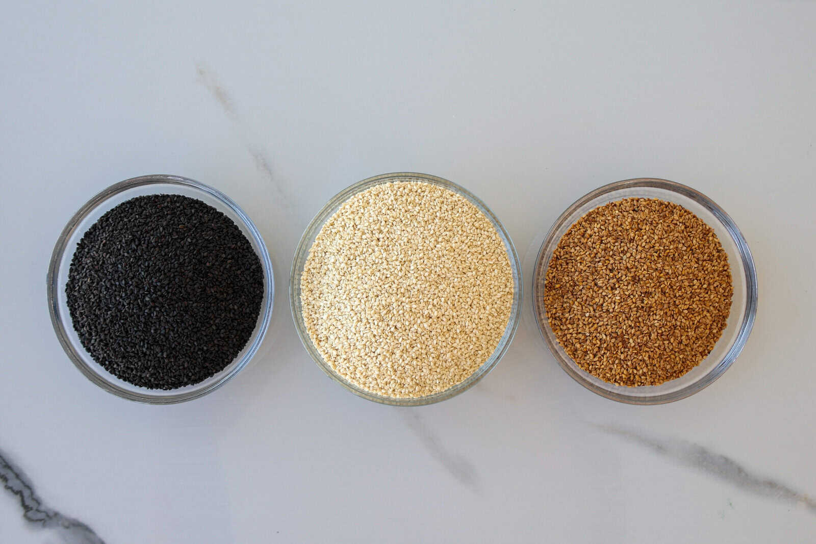 Three small glass bowls are arranged in a row, each filled with different types of seeds: black sesame seeds on the left, white sesame seeds in the middle, and whole sesame seeds on the right, on a light surface.