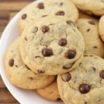 A plate of freshly baked chocolate chip cookies piled on top of each other, showing golden-brown edges and visible chocolate chips.
