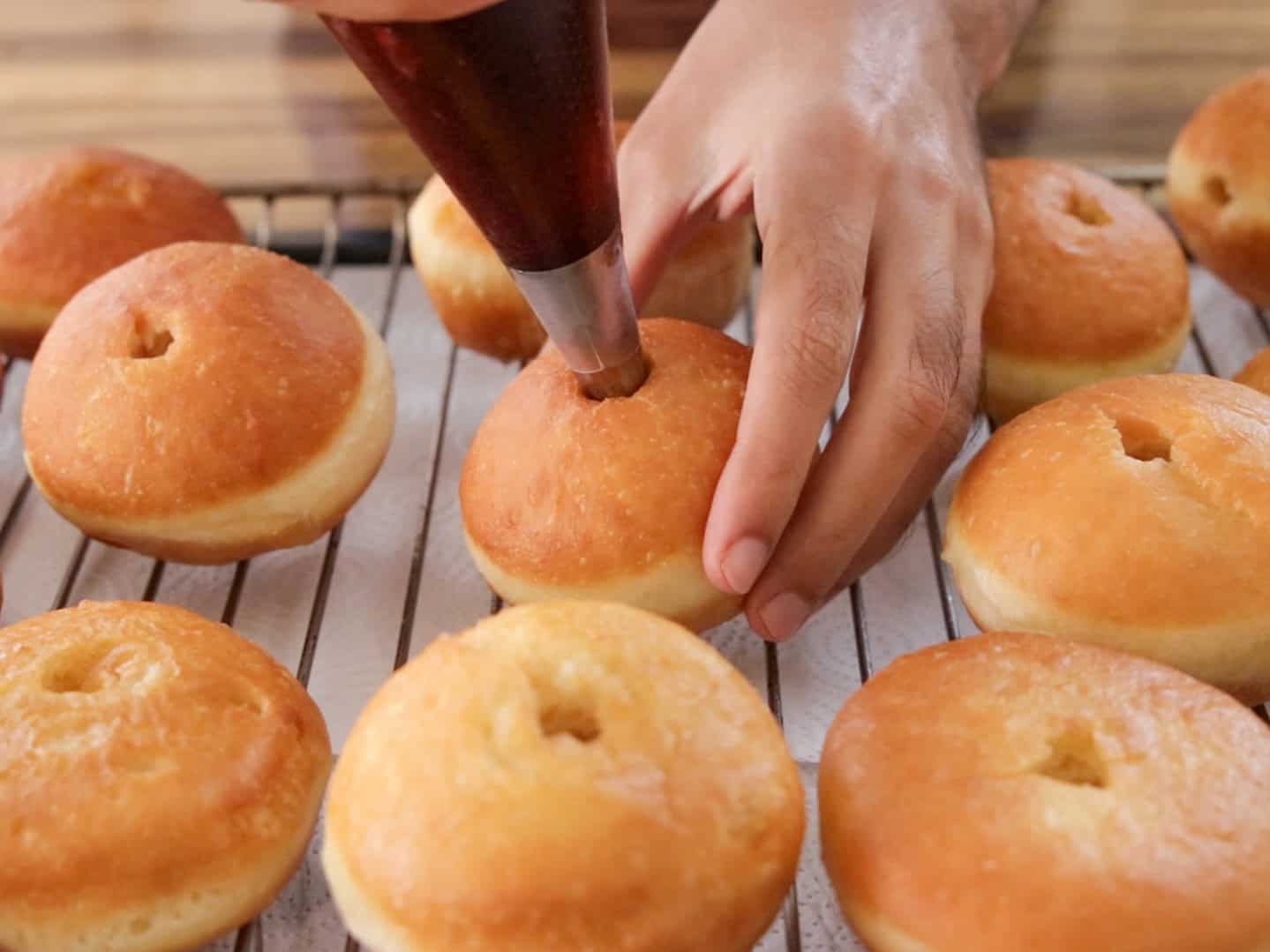 A person fills a round, golden-brown sufganiya doughnut with a piping bag, while several unglazed doughnuts cool on a wire rack lined with paper towels.