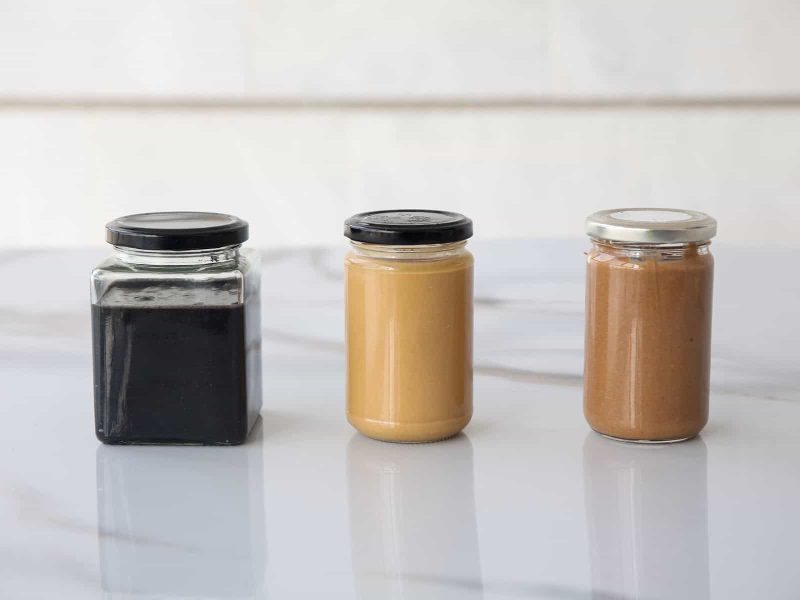 Three jars of spreads on a white marble surface: one with a dark, black spread, one with a light tan spread, and one with a medium brown spread, each with a different style of lid.