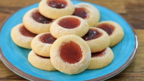 A plate of jam thumbprint cookies with a dusting of powdered sugar and a center filled with red jam, arranged on a turquoise plate atop a wooden surface.