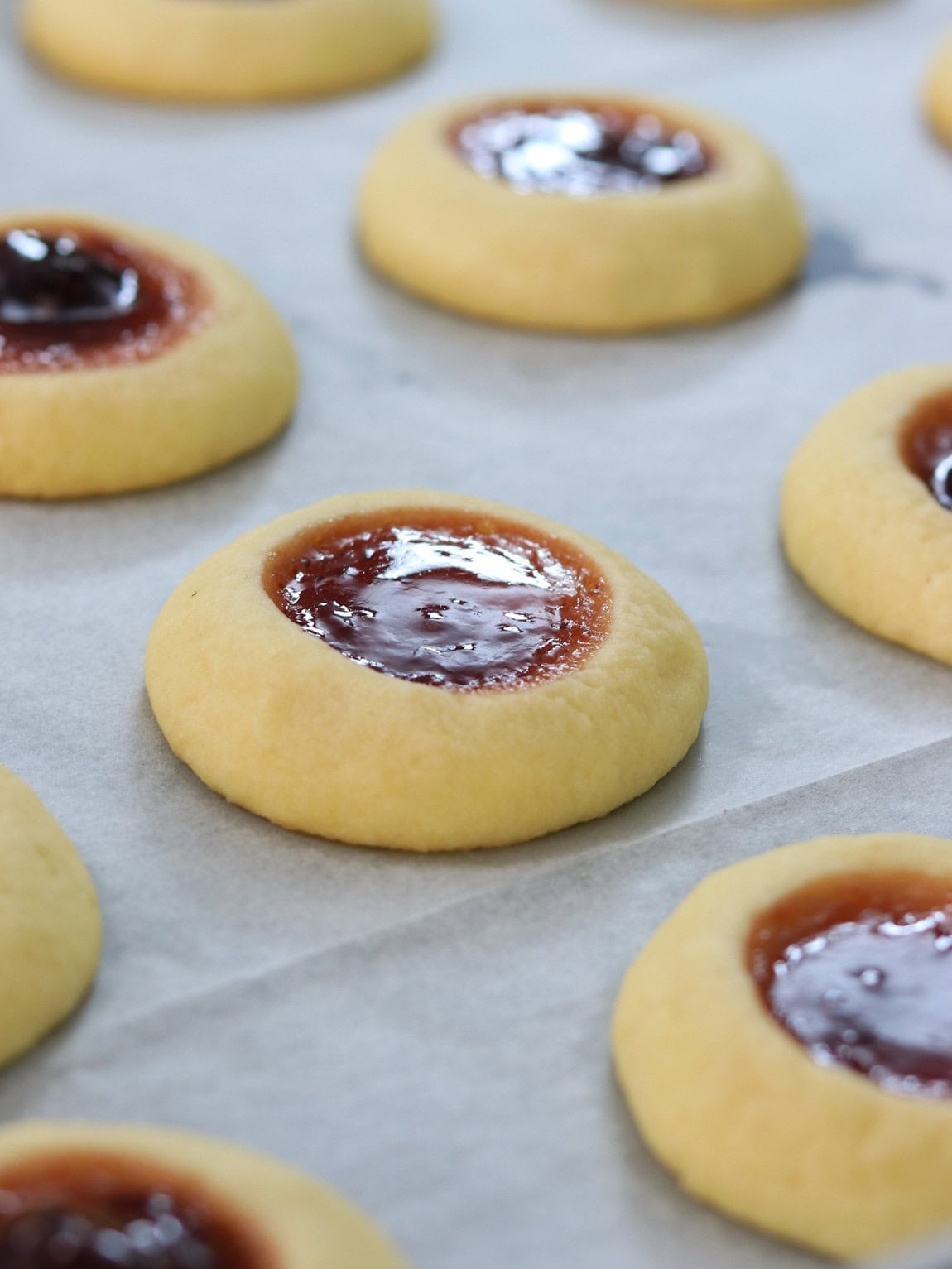 Close-up of freshly baked thumbprint cookies with a glossy red jam filling, arranged on a sheet of parchment paper.