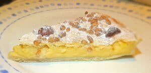 A slice of pie with a creamy yellow filling, topped with powdered sugar and pine nuts, served on a white plate with a blue patterned rim.