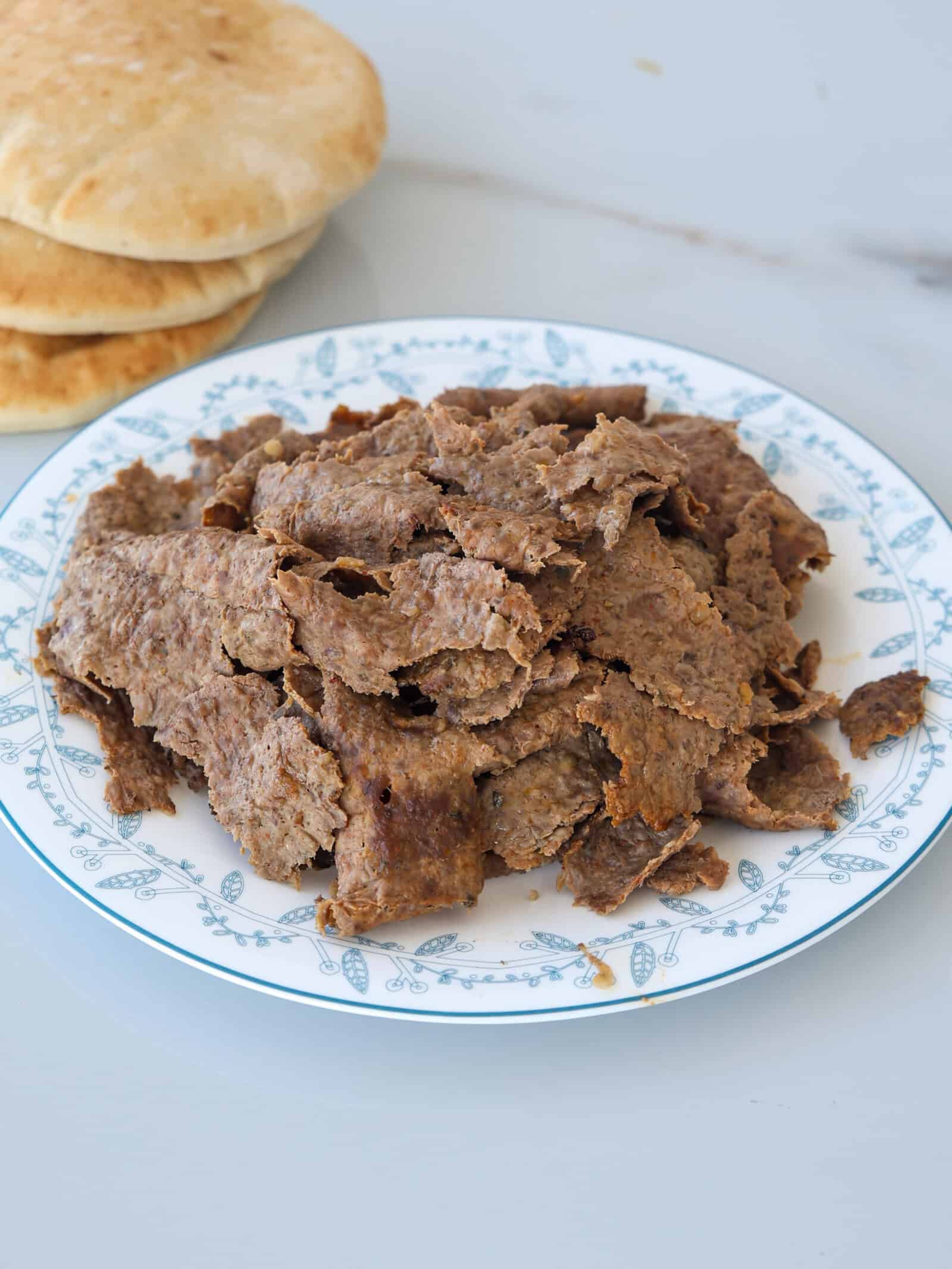 A white plate with sliced doner kebab meat on it, placed on a light surface. In the background, there are several pieces of pita bread stacked on top of each other.